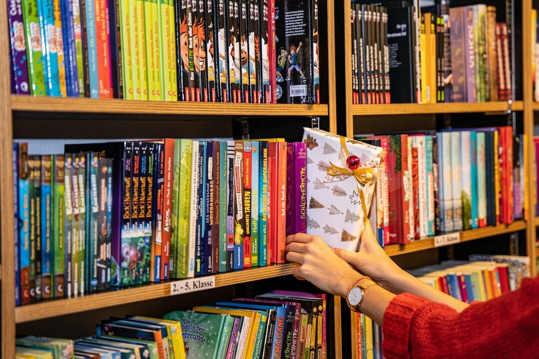 Eine Person stellt ein verpacktes Weihnachtsgeschenk auf ein dicht gefülltes Bücherregal voller bunter Bücher. Das Bücherregal, beschriftet für die Klassen 2 bis 5, bietet Titel, die von skurrilen Geschichten bis zu praktischen Ratgebern wie „Die effektivsten Strategien, um seriös online Geld zu verdienen“ reichen.