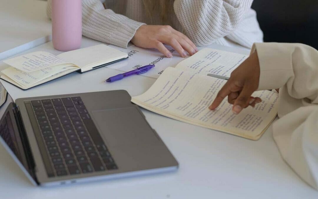 Zwei Personen sitzen mit offenen Notizbüchern, einem Laptop und einem lila Stift an einem Tisch. Eine Person zeigt auf die Notizen im Notizbuch der anderen Person über kostenlose Bücher. Eine rosa Wasserflasche steht ebenfalls auf dem Tisch.